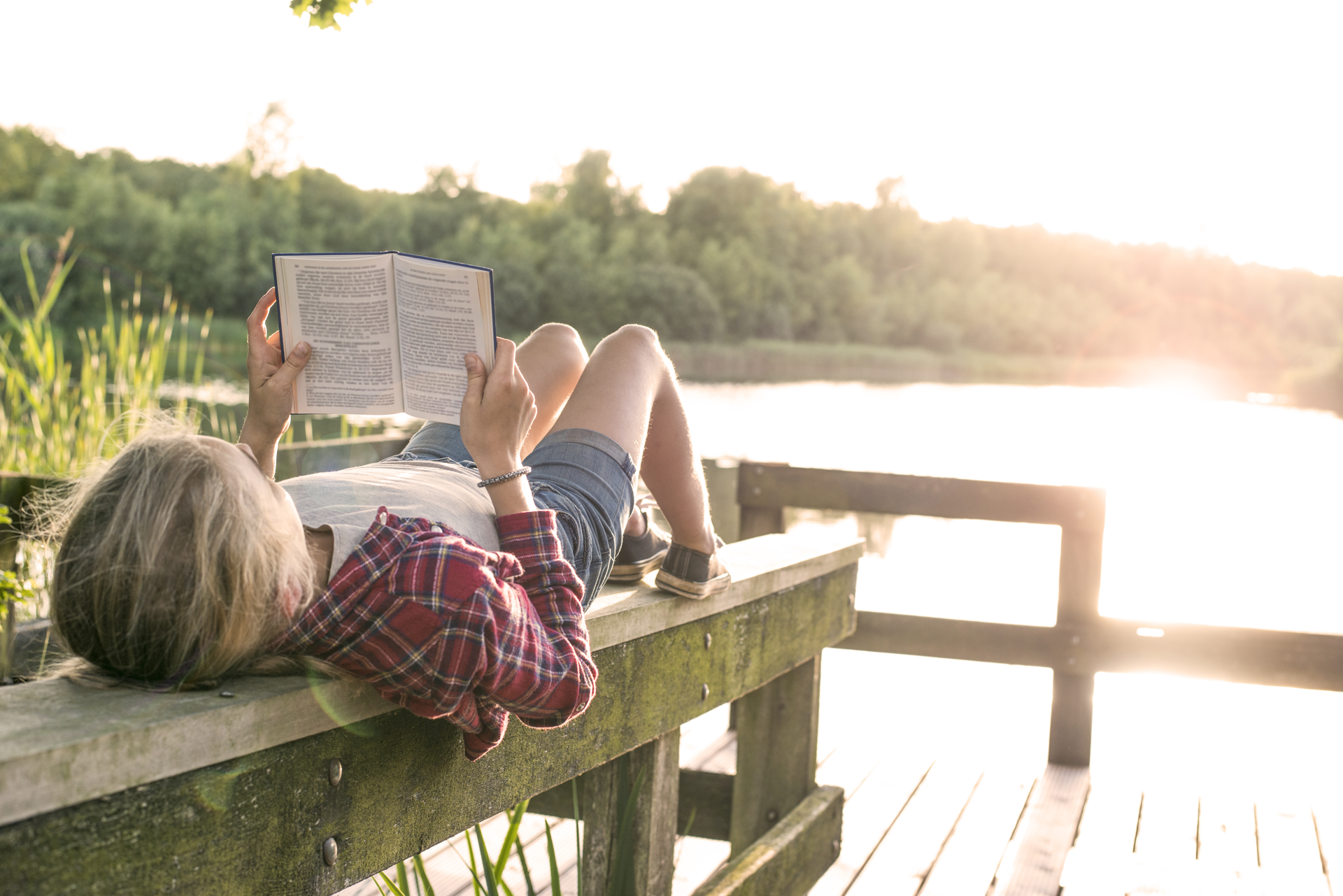 Ein Mädchen liest ein Buch draußen in der Sommersonne am See.