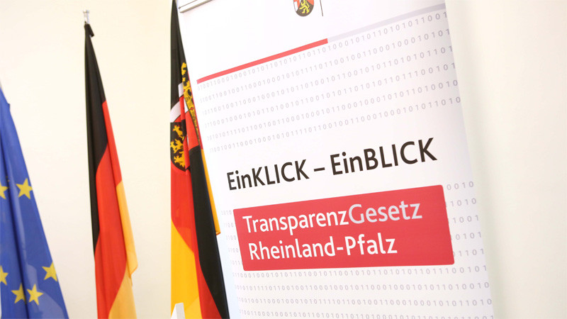 Drei Fahnen von Europa, Deutschland und Rheinland-Pfalz stehen vor einem Plakat. Darauf ist zu lesen: EinKLOCK - EinBLICK. TransparenzGesetz Rheinland-Pfalz