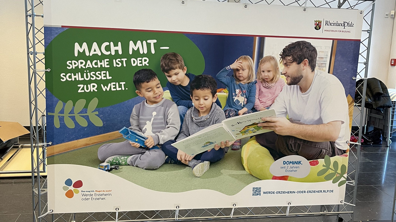 Zu sehen ist ein Plakat der Kampagne. Darauf abgebildet ein Erzieher, der gemeinsam mit fünf Kindern ein Buch liest. Oben ist eine Sprechblase abgebildet auf der steht: Macht mit- Sprache ist der Schlüssel zur Welt