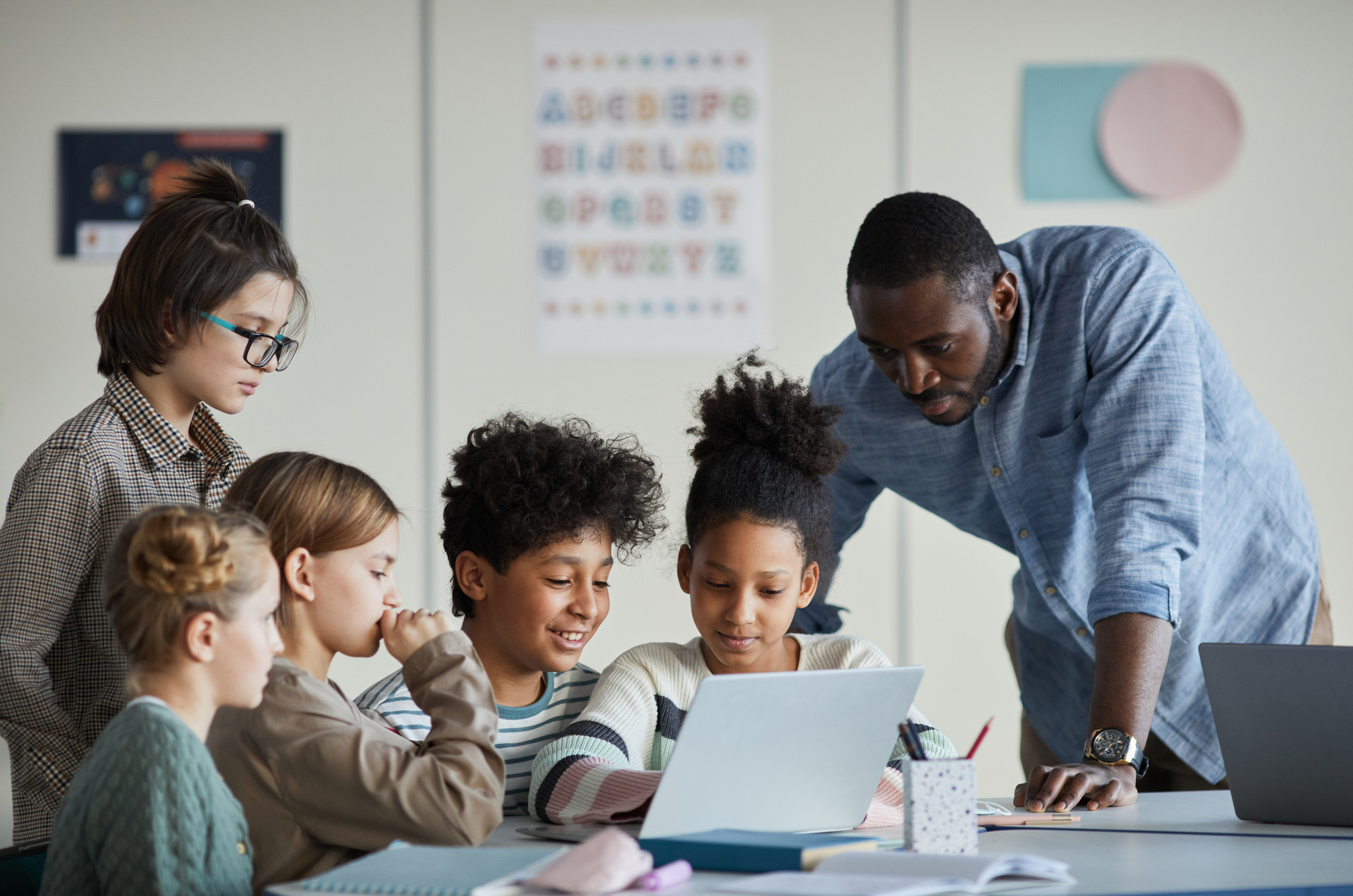 Mehrere Kinder sitzen um ein Tablet herum. Ein Lehrer erklärt den Kindern den Umgang.