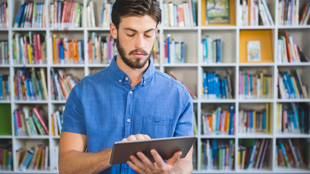 Ein junger Mann lernt am Tablet, er steht vor einem Bücherregal