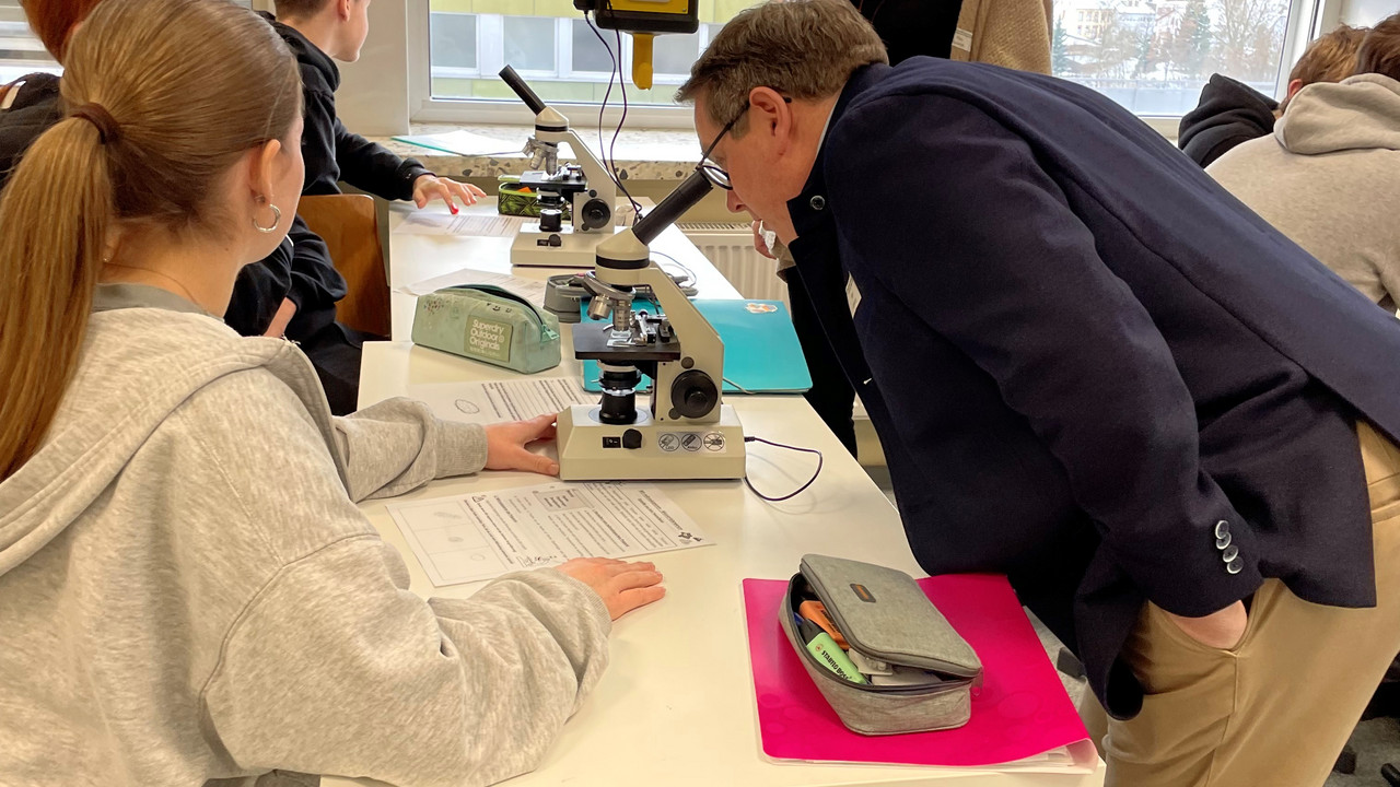 Auf dem Bild ist ADD-Präsident Linnertz mit Schülerinnen und Schülern während des Biologieunterrichts zu sehen. Linnertz schaut durch ein Mikroskop. 