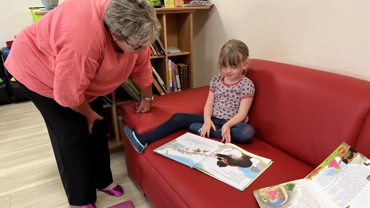 Staatsekretärin Brück bückt sich herunter zu einem Mädchen, was auf einer Couch sitzt und ein Buch vor sich hat. Sie unterhalten sich über das Buch. 