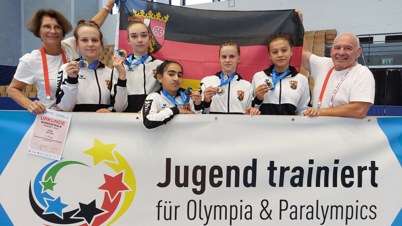 Zu sehen sind die fünf Mädchen sowie ihre Betreuer. Sie halten alle stolz ihre Medaillen in die Kamera. Im Hintergrund wird die Landesflagge hoch gehalten. 