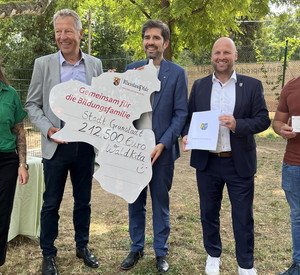 Auf dem Foto sind links nach rechts die stellvertretende Kita-Leitung Vivian Heim, Bürgermeister Klaus Wagner, Bildungsminister Sven Teuber, Erster Kreisbeigeordneter Timo Jordan sowie Kita-Leiter Markus Schubert zu sehen. Sie halten gemeinsam den Förderbescheid in Form des Landes Rheinland-Pfalz. 