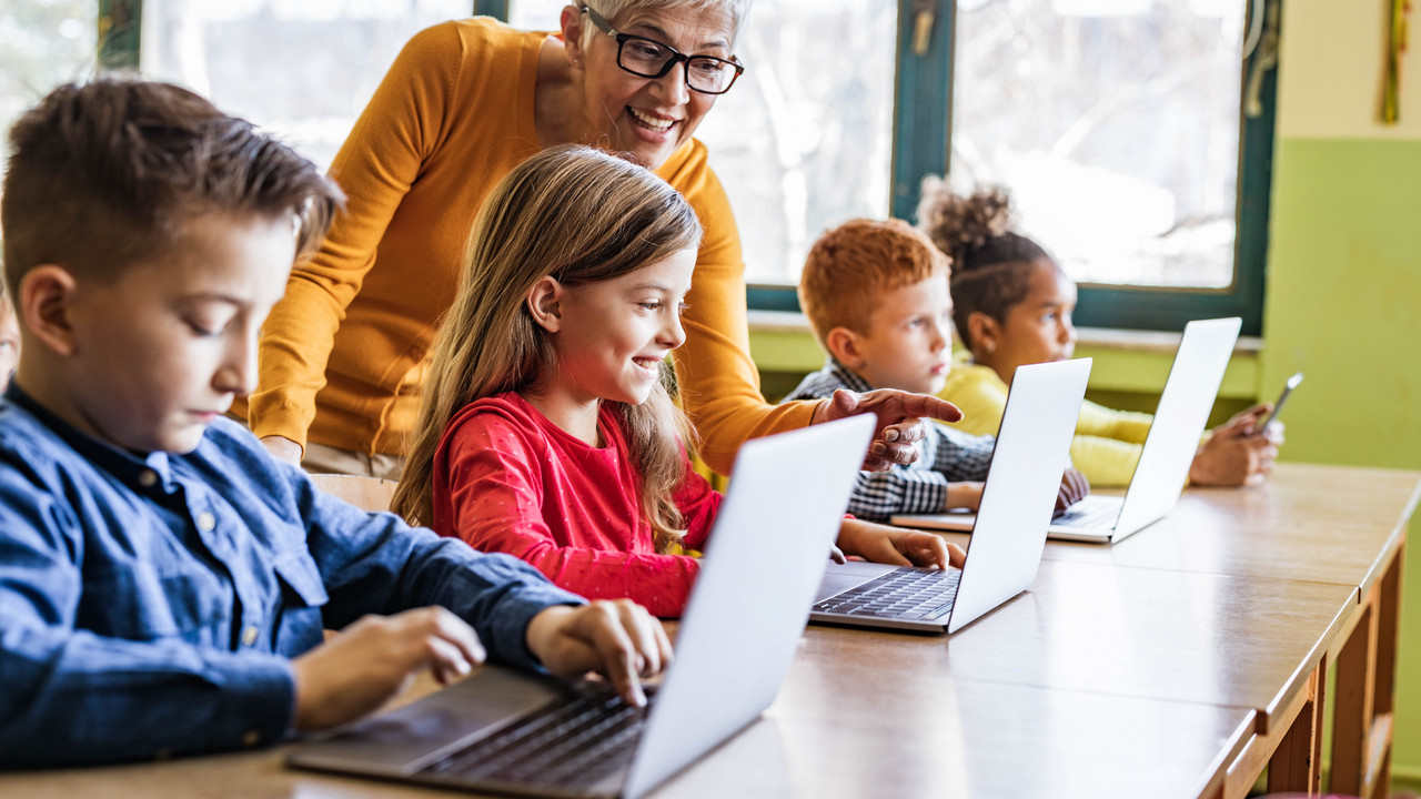 Man sieht eine Lehrerin, die ihre Schülerinnen und Schüler bei der Arbeit am Laptop unterstützt