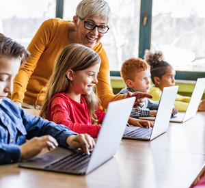 Man sieht eine Lehrerin, die ihre Schülerinnen und Schüler bei der Arbeit am Laptop unterstützt