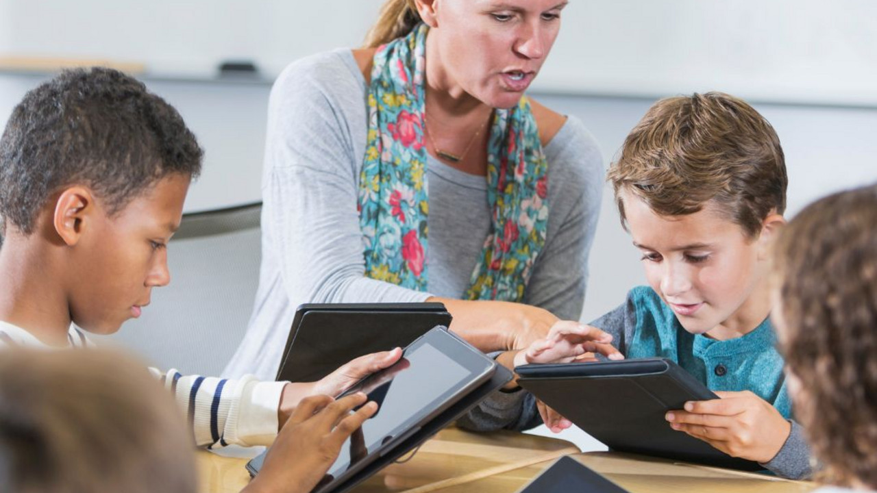 Auf dem Bild sieht man Schülerinnen und Schüler die an Tablets arbeiten. Sie werden von einer Lehrkraft angeleitet.