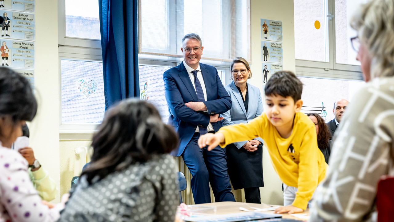 Ministerpräsident Alexander Schweitzer und Bildungsministerin Dr. Stefanie Hubig stehen im Hintergrund und beobachten die Arbeit der Lehrerin mit den Kindern an der Goethe-Schule.