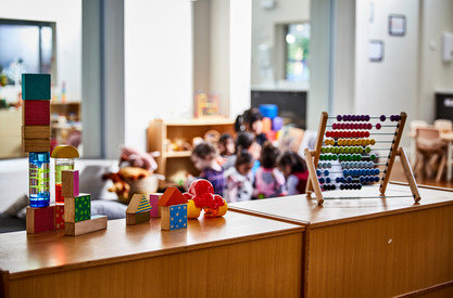 Im Vordergrund: Spielzeuge auf einem Tisch, Im Hintergrund: Kinder spielen im Kindergarten