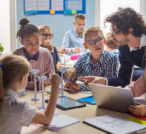 Gruppe Schülerinnen und Schüler sitzen am Tisch. Auf dem Tisch stehen Modell-Windräder. Einige arbeiten mit Tablets. Ein Lehrer beugt sich zu Ihnen und erklärt etwas. 