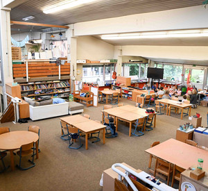 Auf dem Foto ist ein offen gestalteter Lernraum zu sehen. Es gibt mehrere Gruppentische, im Hintergrund sitzt eine Gruppe aus Schülerinnen und Schülern im Kreis auf dem Boden. 